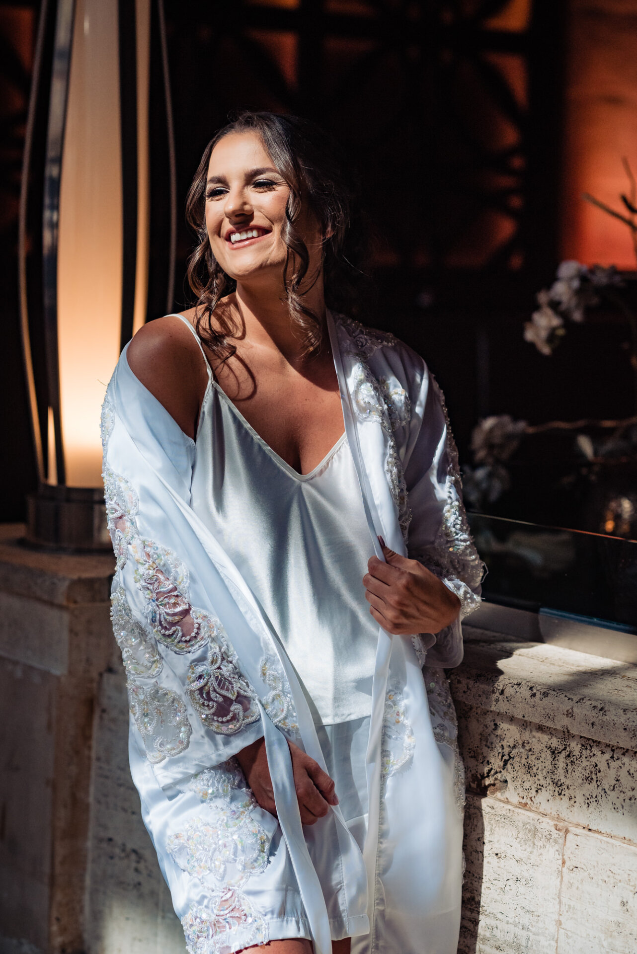 Radiant and filled with anticipation, the woman smiles as she stands in her white satin robe, moments before her wedding.