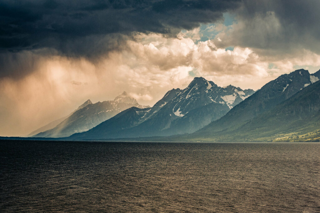 A dramatic scene unfolds as a storm passes by the Grand Tetons, with dark clouds swirling overhead and the tranquil lake below reflecting the dramatic sky, capturing the raw power and beauty of nature in Wyoming's iconic mountain range