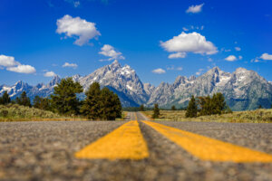 A stunning landscape shot captures the majestic Tetons mountain range rising in the distance, framed by the paved road leading through the scenic wilderness of Wyoming