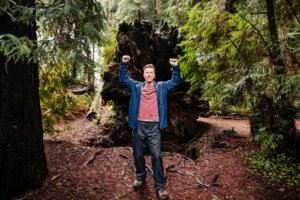 man is standing behind a towering tree that has fallen. This once magnificent redwood tree, standing at an impressive 20 feet in diameter, now lies on the forest floor, its massive trunk and sprawling branches a testament to the grandeur of nature.