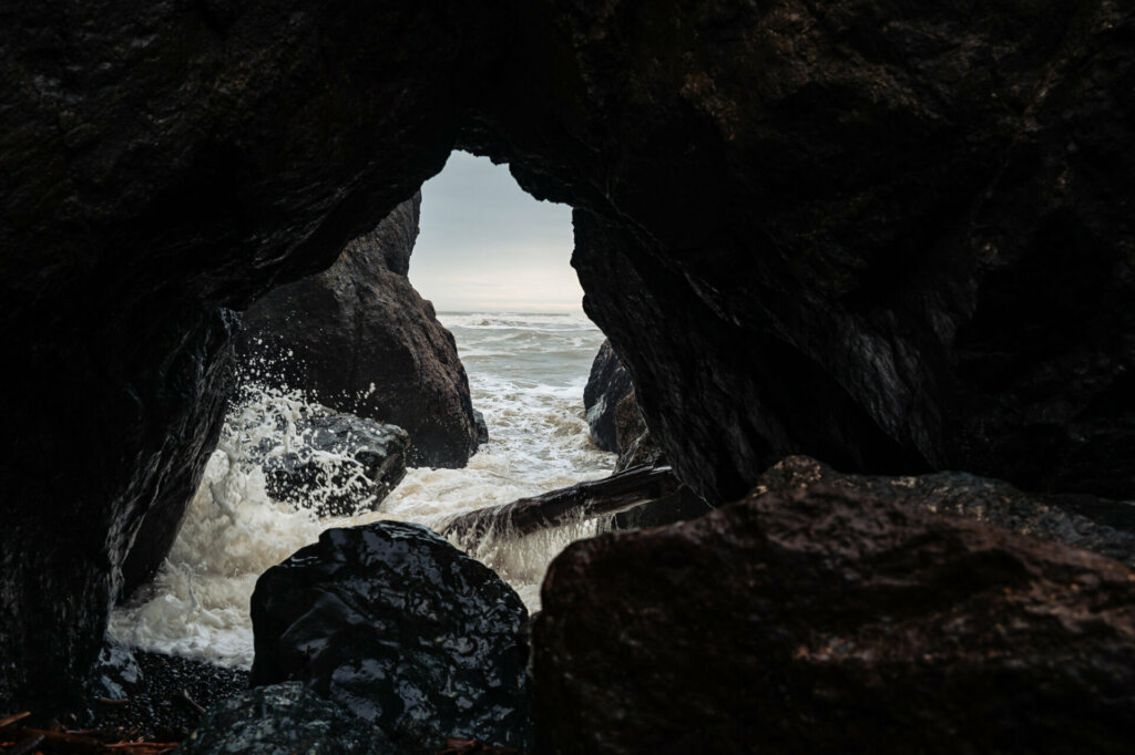 A small rock cave nestled along the shoreline of Kalaloch Beach, framed by towering cliffs and battered by the relentless force of huge waves crashing against the rugged coastline.