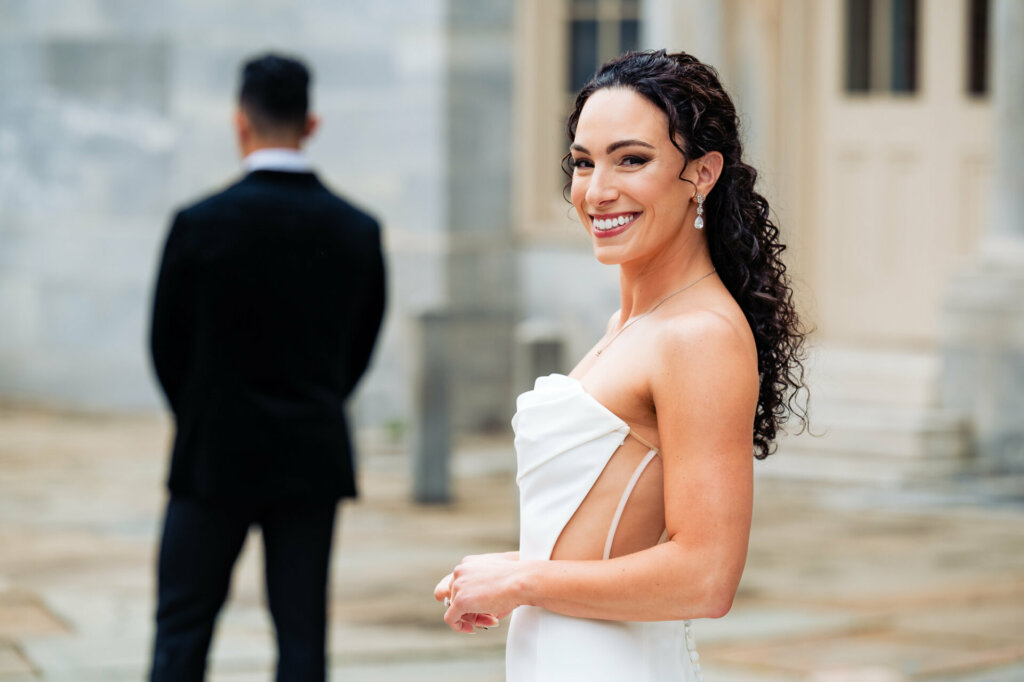 Bride smiles for the camera in anticipation of the first look, while the groom waits with eager anticipation, facing away