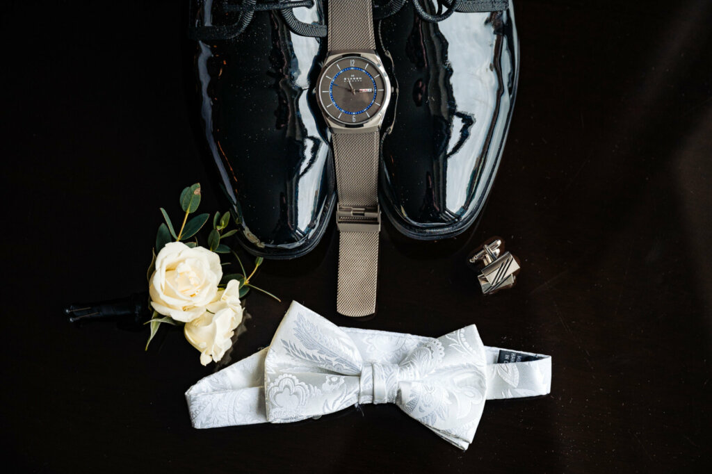 Groom's wedding attire featuring a white ribbon tie, formal shoes, watch, and other accessories laid out for display.