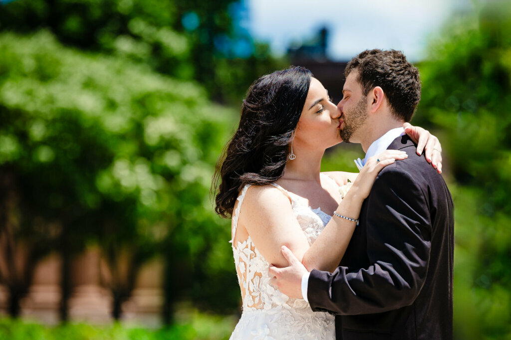 Close-up of bride in white lace gown and veil kissing groom in dark suit. Couple embracing tenderly against a soft-focus background of green trees and foliage. Romantic wedding portrait capturing intimate moment.