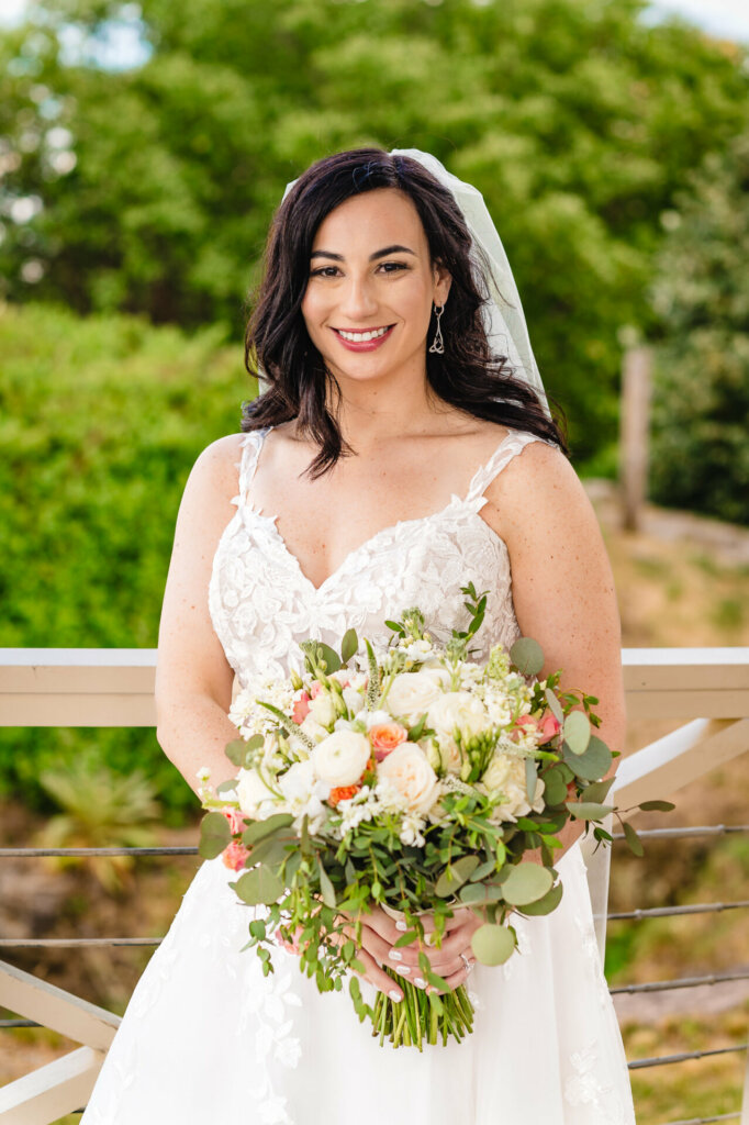 Close-up of radiant bride with natural makeup, wearing white gown and veil, holding bouquet against blurred nature backdrop