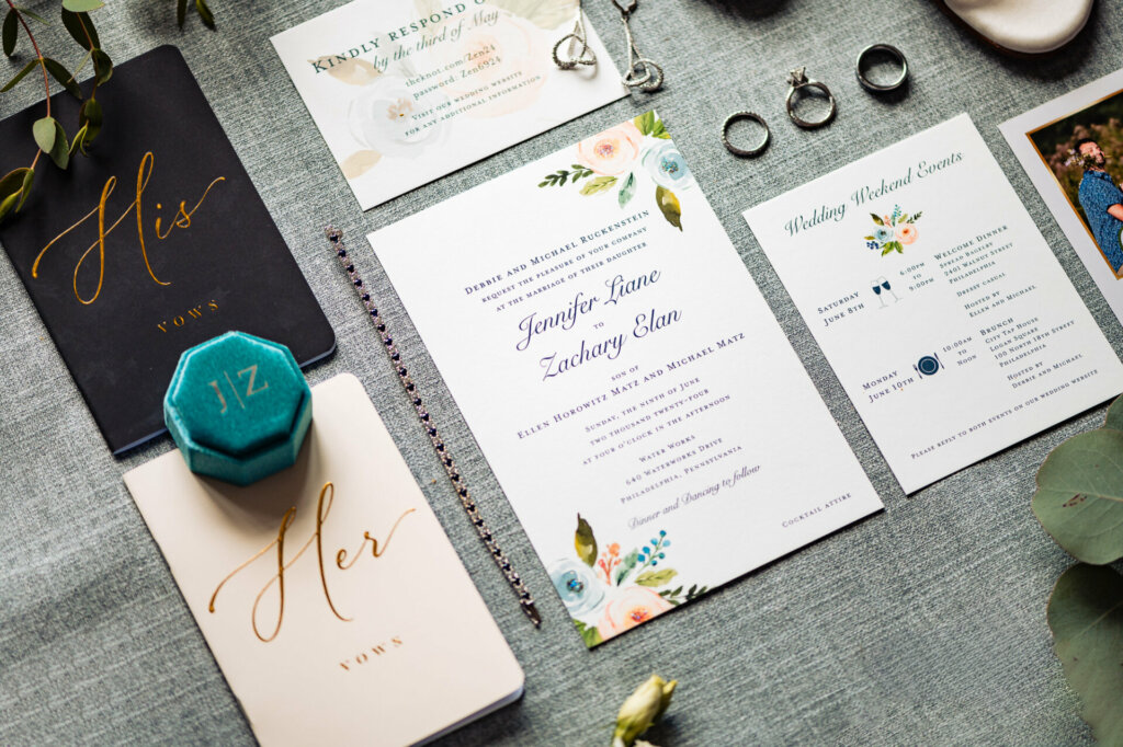Wedding stationery ensemble featuring handwritten vows, personal letters, bride's accessories including jewelry and heels, and an invitation laid out on a gray background with floral accents.