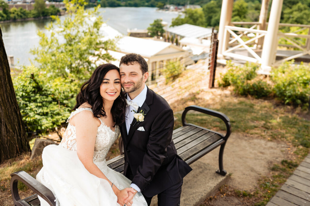 Newlywed couple in wedding attire sitting on a bench, holding hands and smiling at the camera.