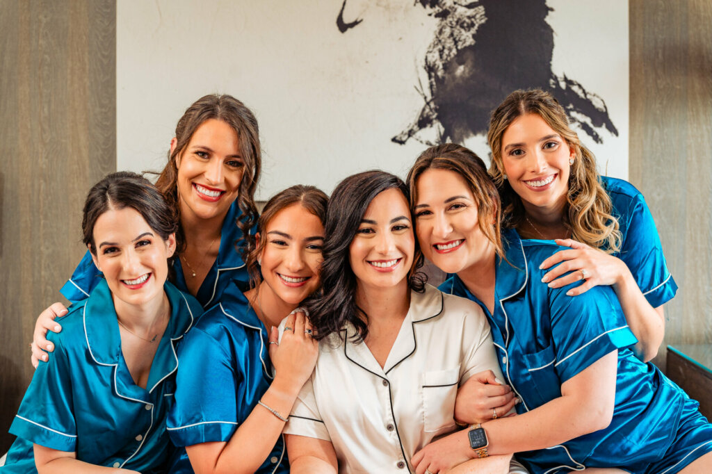 Joyful bride in white silk pajamas surrounded by her bridesmaids in blue silk pajamas, all smiling together.