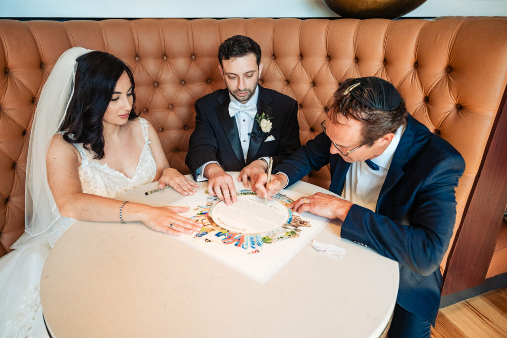 Bride and groom attentively review and sign the ketubah, their Jewish marriage contract, witnessed by loved ones.