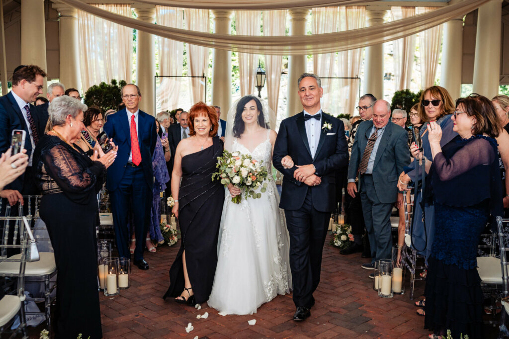 Proud parents, beaming with joy, escort their radiant daughter down the aisle to begin her new chapter as a wife.