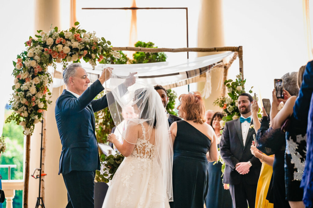 A tender moment unfolds as the bride's father adjusts her veil, a silent exchange of love and pride before giving her hand in marriage.