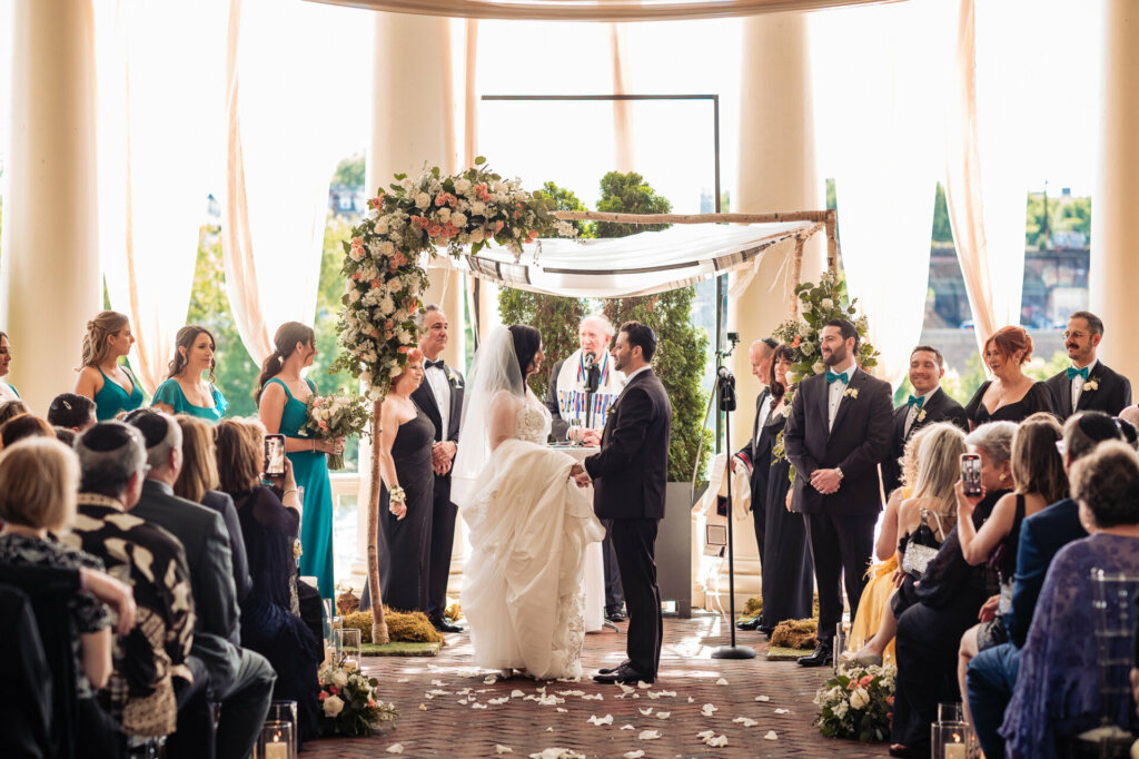 Under the open canopy of a chuppah, a radiant bride and groom exchange vows in a heartfelt Jewish wedding ceremony.