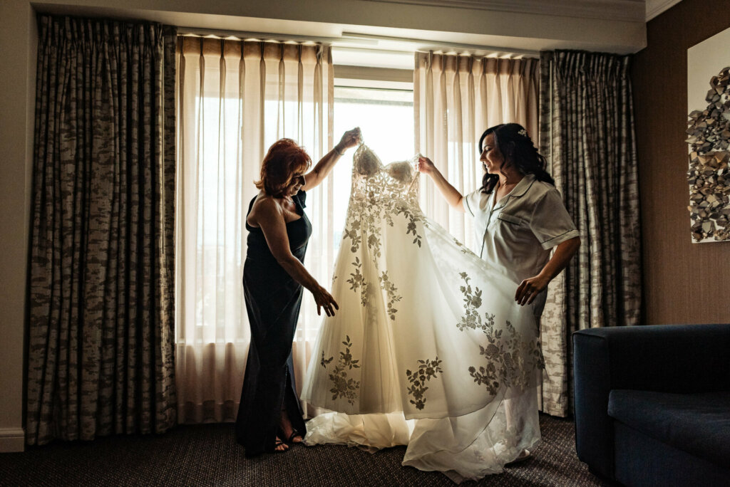Bride and her mother smiling joyfully as they hold up the wedding gown, admiring it together.