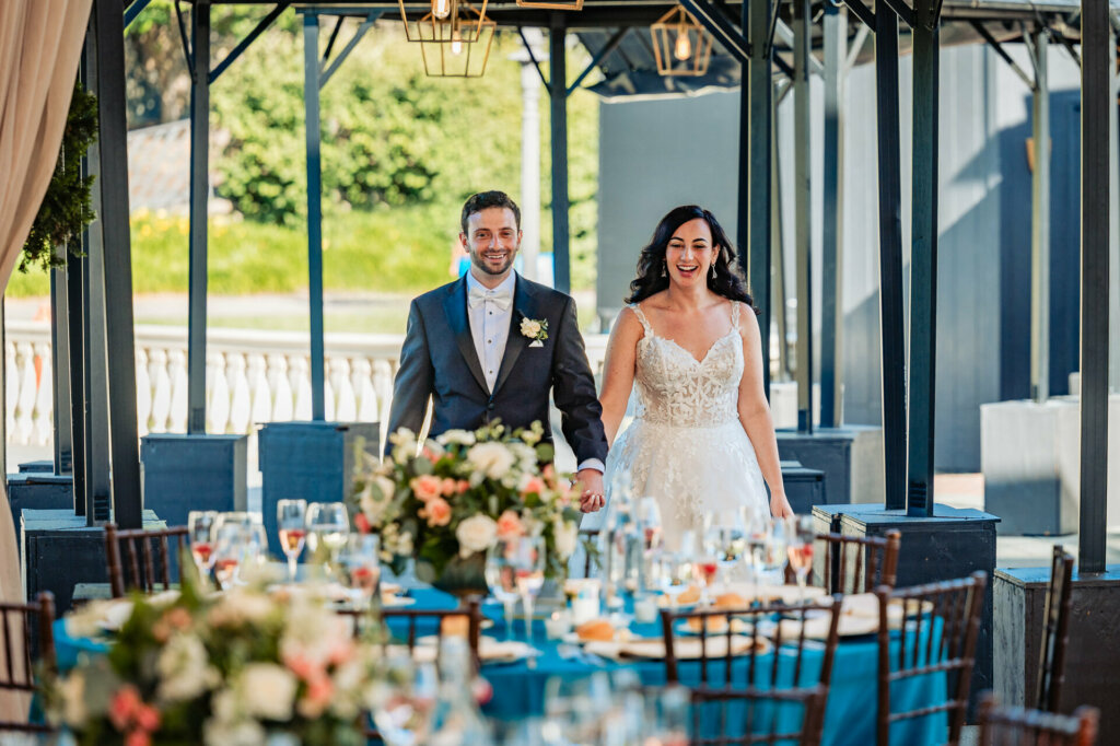 Joyful newlyweds entering their wedding reception hand-in-hand, faces lit up with smiles as they take in the scene.