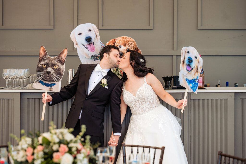 Newlyweds sharing a kiss while holding hands and displaying cut-out pictures of their pets during their wedding reception.