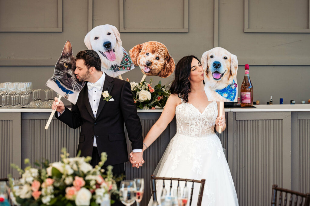 Newlyweds posing with a kiss while holding hands and displaying cut-out pictures of their pets during their wedding reception.