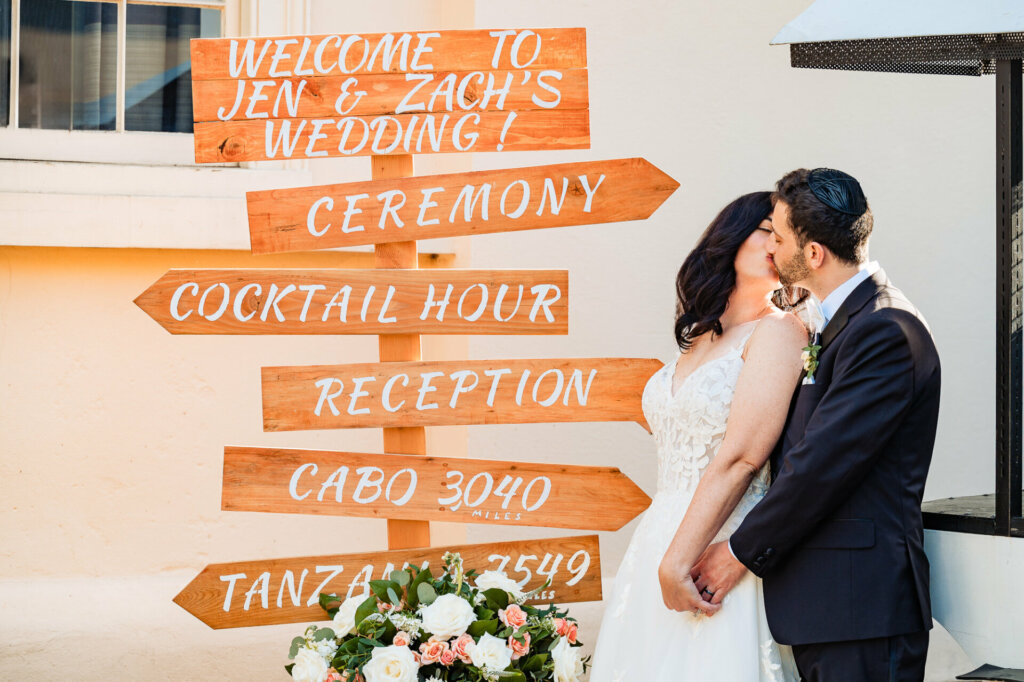Bride and groom sharing a kiss beside their wedding reception decor, featuring decorative signs.