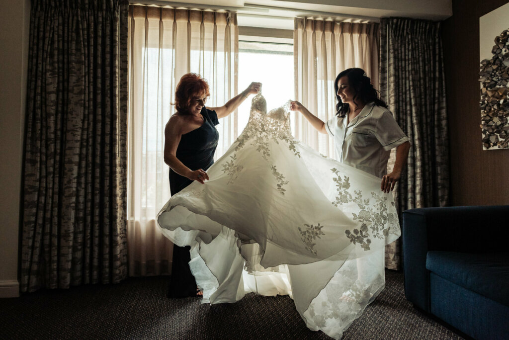 Bride and her mother smiling joyfully as they hold up the wedding gown, admiring it together.