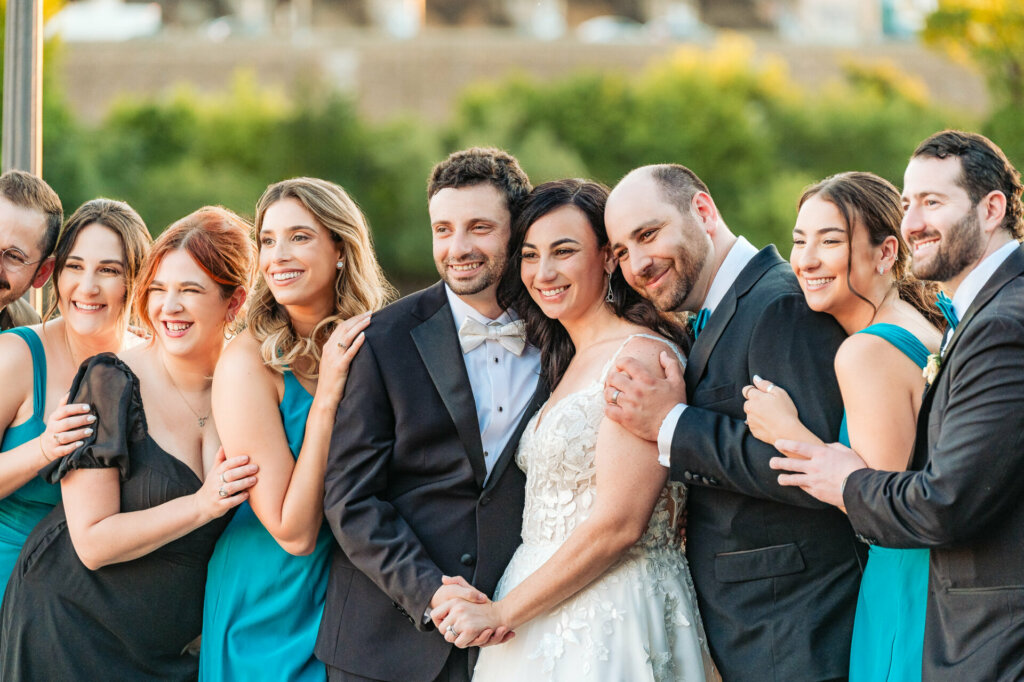 Jubilant newlyweds, radiant in their wedding attire, celebrate with their bridesmaids and groomsmen on a scenic lakeside terrace.
