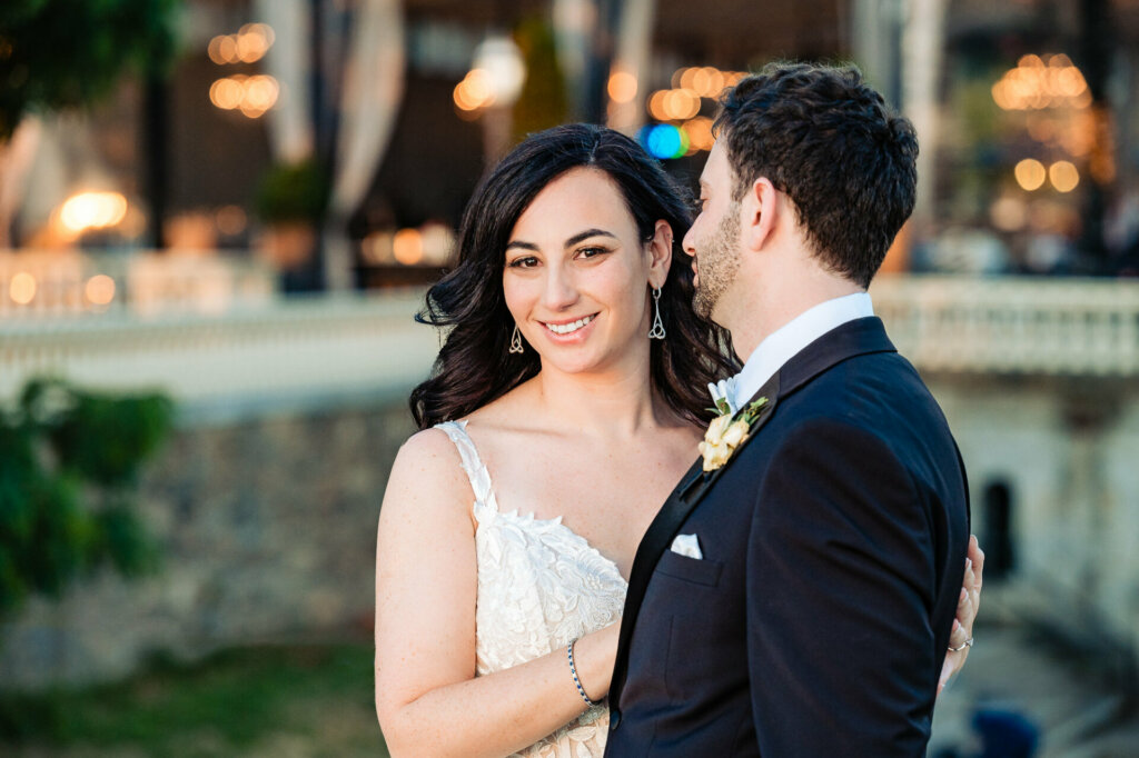 Radiant bride beams for the camera, bathed in the groom's adoring gaze, on their wedding day.