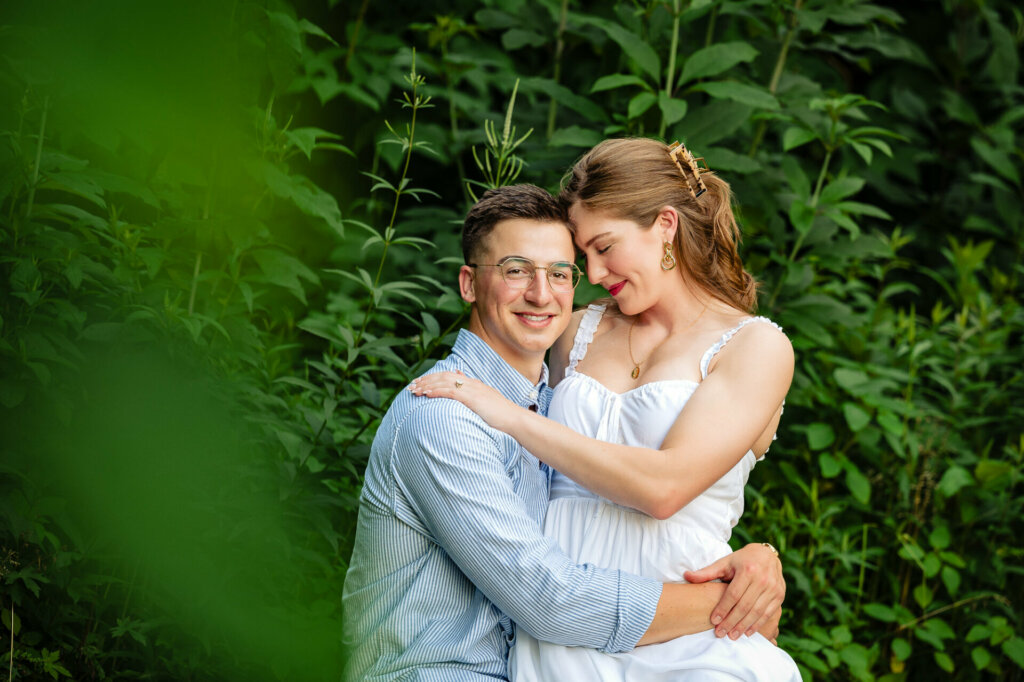 A smiling couple in a lush green setting. The man wears glasses and a blue striped shirt. The woman has her hair up and wears a white dress. They are embracing, with the woman's head resting against the man's.