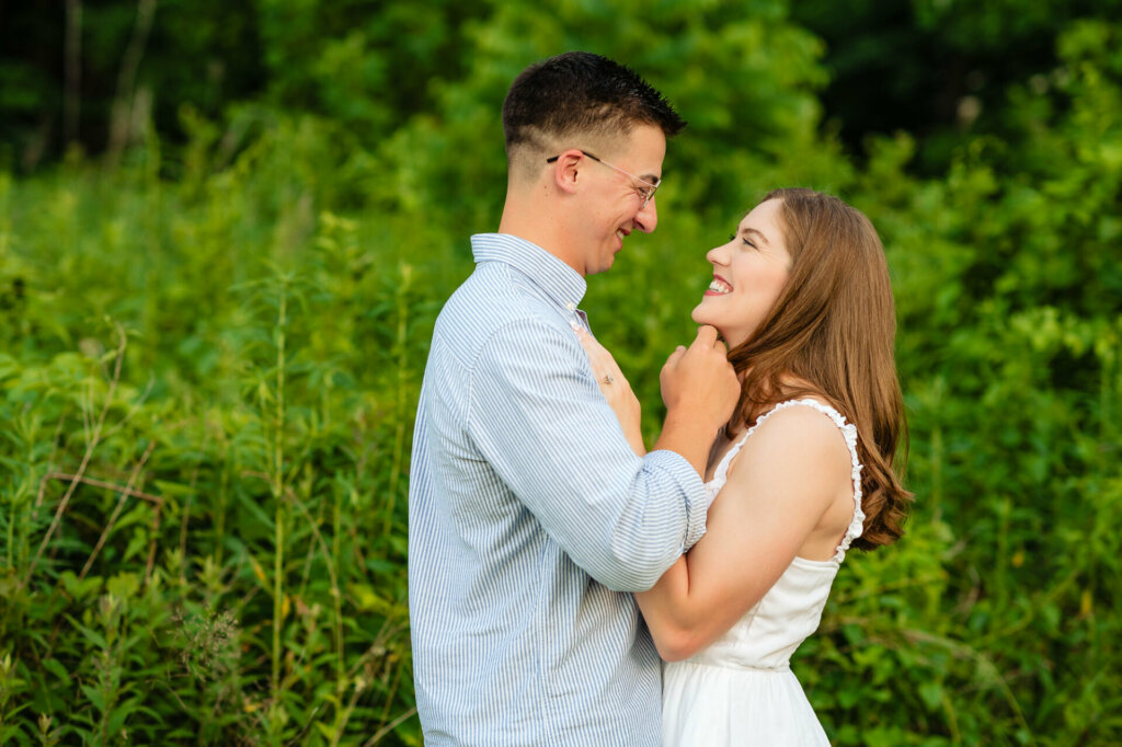 A young couple, standing close together in a green field, gazing lovingly into each other's eyes. The man is wearing a light blue striped shirt and glasses, while the woman is in a white dress. The lush greenery in the background complements their intimate pose, creating a serene and romantic atmosphere.