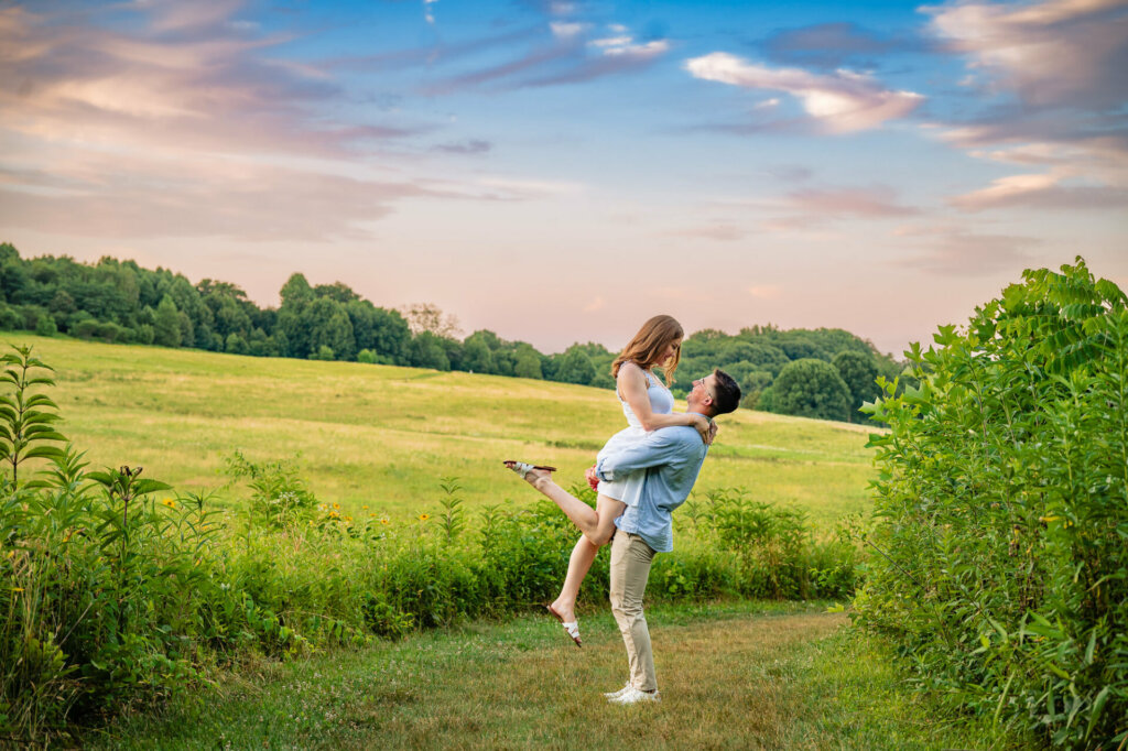This image shows a romantic scene of a couple in a field at sunset. The man is lifting the woman, who has her arms around his neck. They are surrounded by lush green vegetation with a golden field and tree line visible in the background. The sky has a warm, pastel glow with soft clouds.