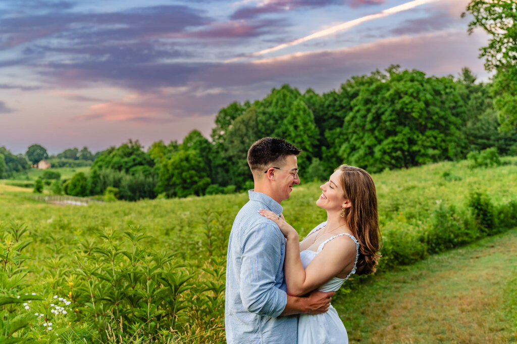 A couple, embraced and smiling at each other, standing in a vast green field during sunset. The man is in a light blue striped shirt and glasses, while the woman wears a white dress. The background features a mix of green trees and a colorful sky with pink and purple hues, enhancing the romantic mood of the scene.