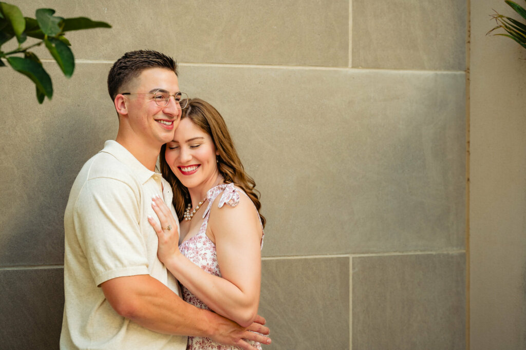 Intimate portrait of the couple against a stone wall. The man in a white shirt hugs the woman in a pink floral dress from behind. Both are smiling warmly, with the woman's head resting on the man's shoulder. Green foliage is visible at the edges.