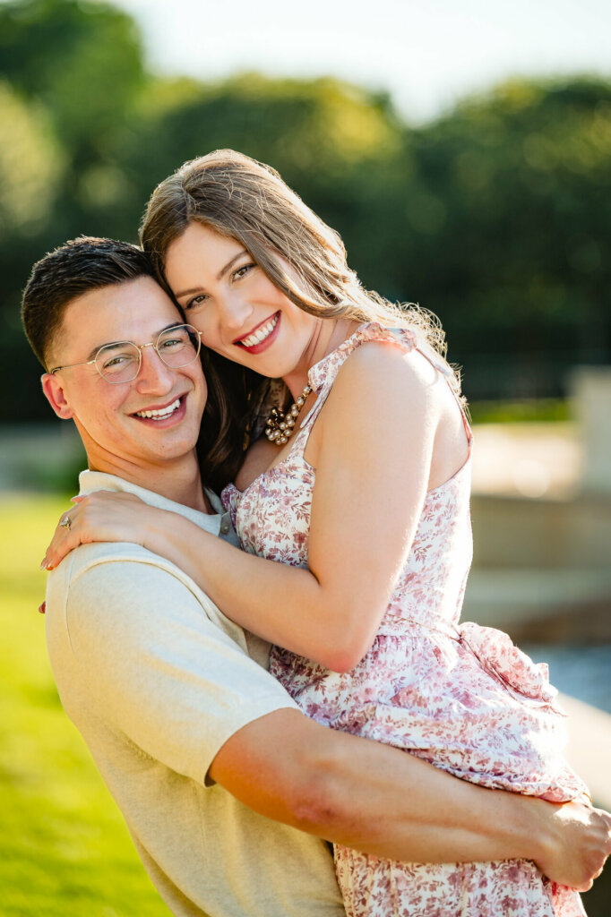 Close-up portrait of the happy couple outdoors. The man has short dark hair and glasses, wearing a light-colored shirt. The woman has long wavy hair and a floral top. They're embracing tightly, with bright smiles and sun-lit greenery in the background.