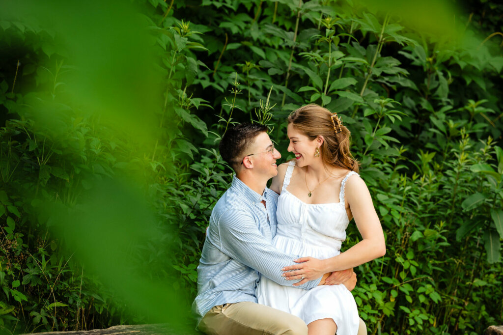 A couple sitting together in a verdant outdoor setting. The man wears glasses and a blue striped shirt. The woman has her hair partially up and wears a white dress. They are looking at each other affectionately, surrounded by lush green foliage.