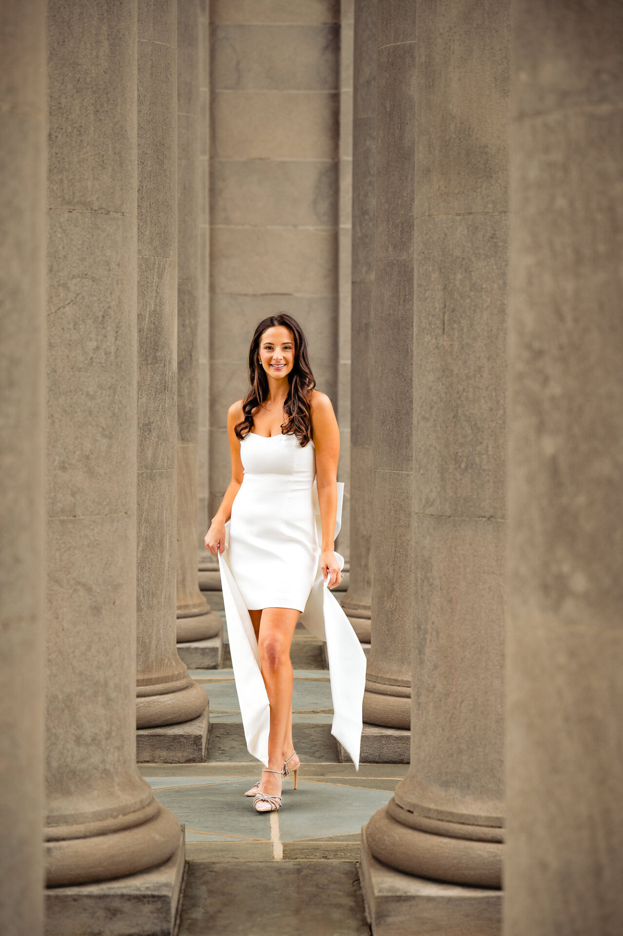 A woman wearing a white, figure-hugging dress smiles warmly at the camera during her engagement session.