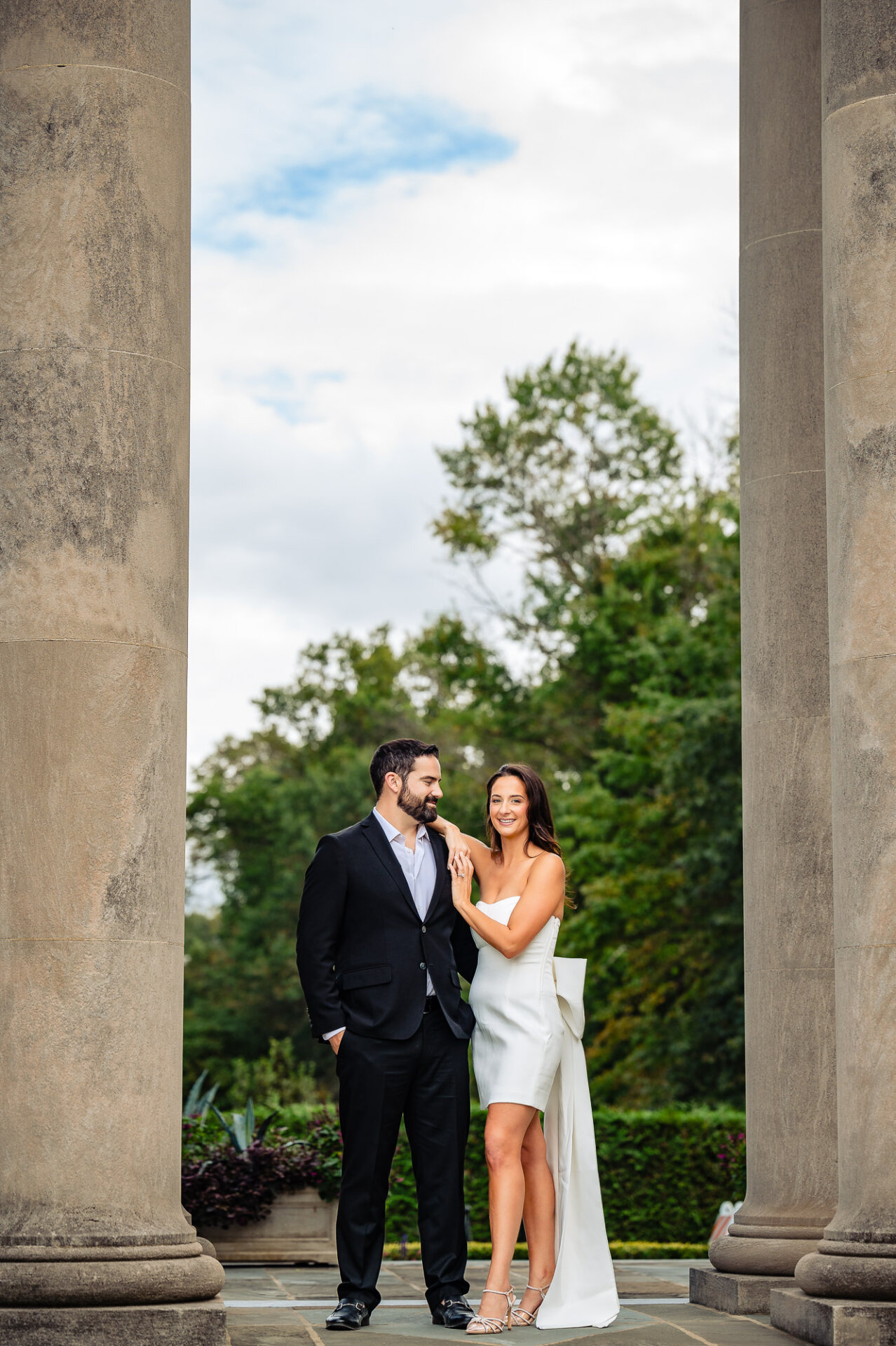 An affectionate couple standing close in an outdoor setting, the man embracing his partner while they look directly into the camera.