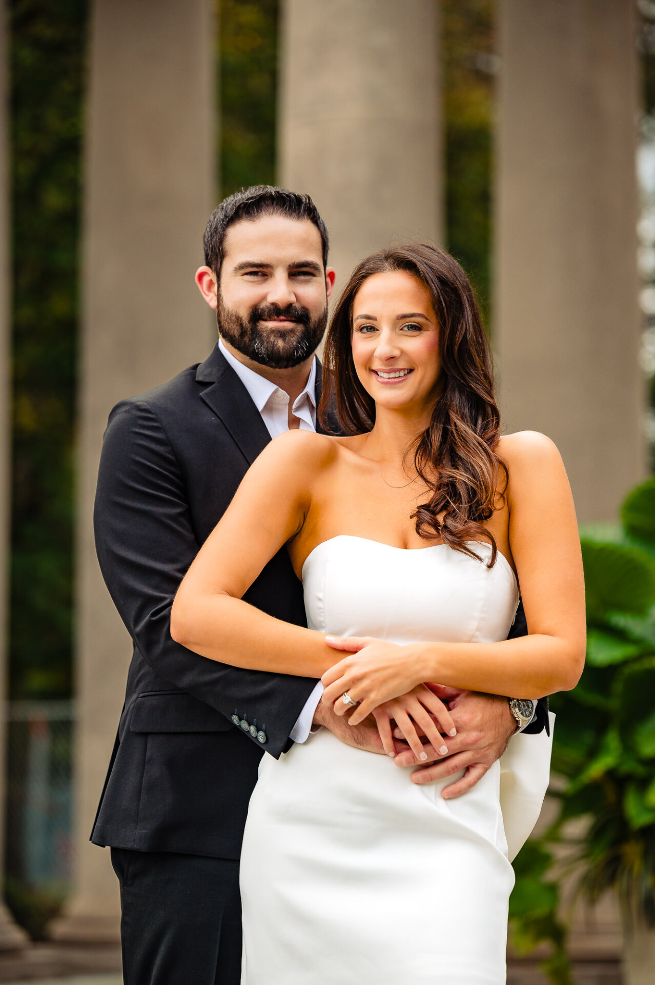 An affectionate couple standing close in an outdoor setting, the man embracing his partner while they look directly into the camera.