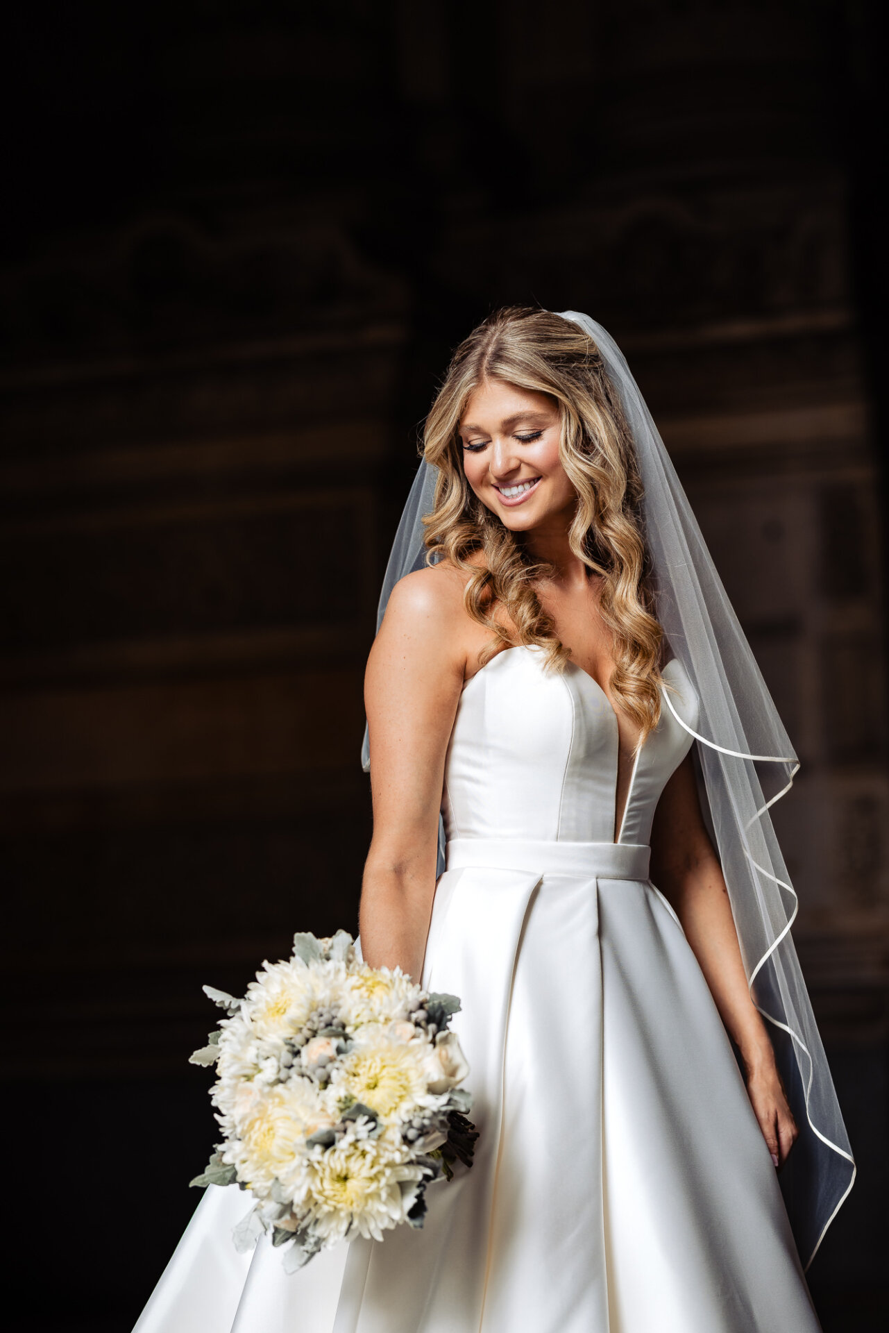 Bridal portrait of the bride smiling gently while looking down, wearing her veil and holding a her wedding bouquet.