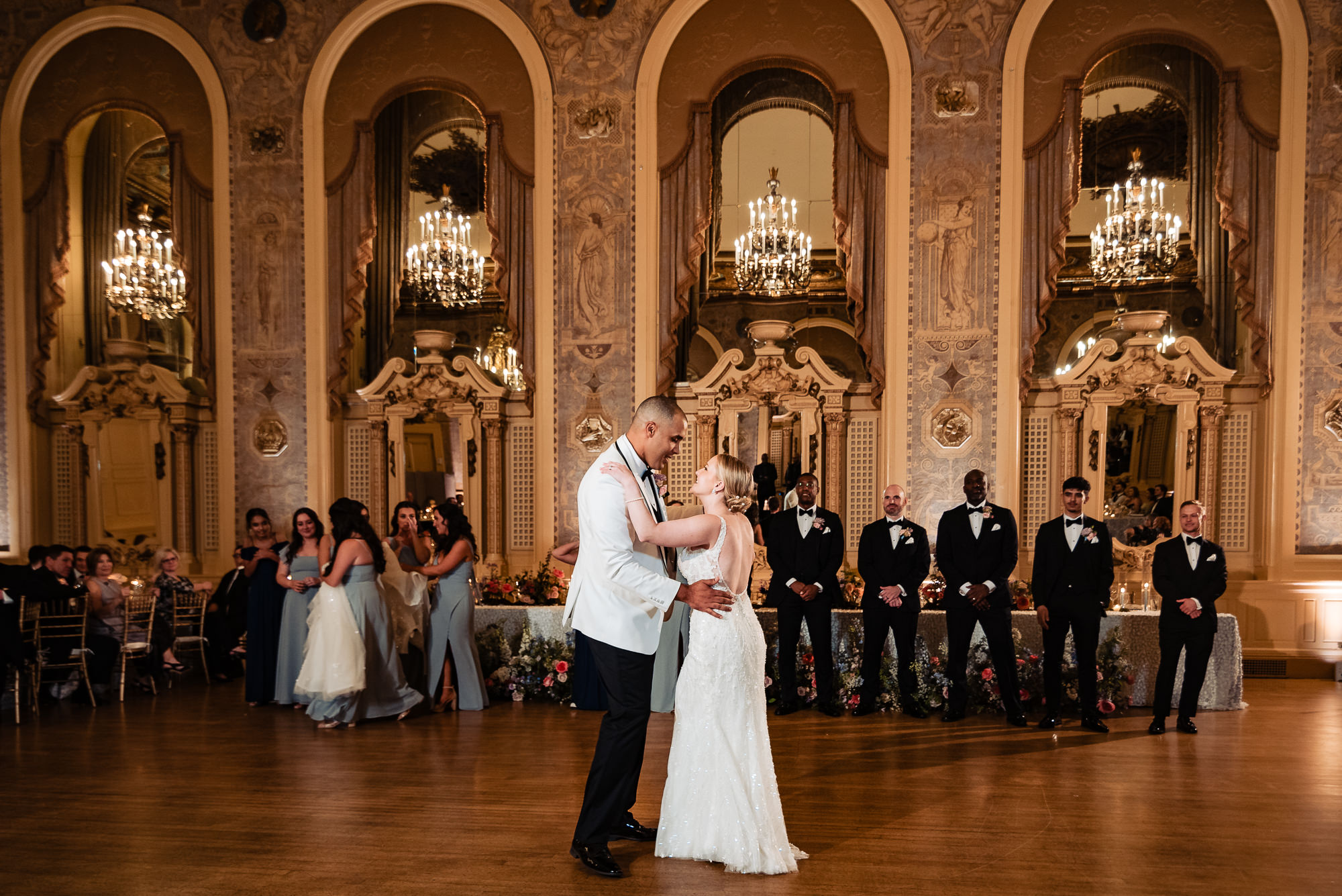 Bride and groom sharing their first dance in a grand ballroom, with bridesmaids and groomsmen watching in the background