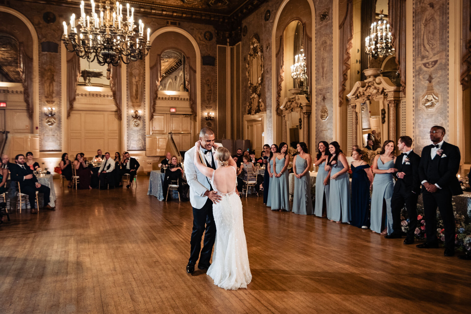 Bride and groom mid-dance, smiling at each other as wedding guests and bridal party watch in the background