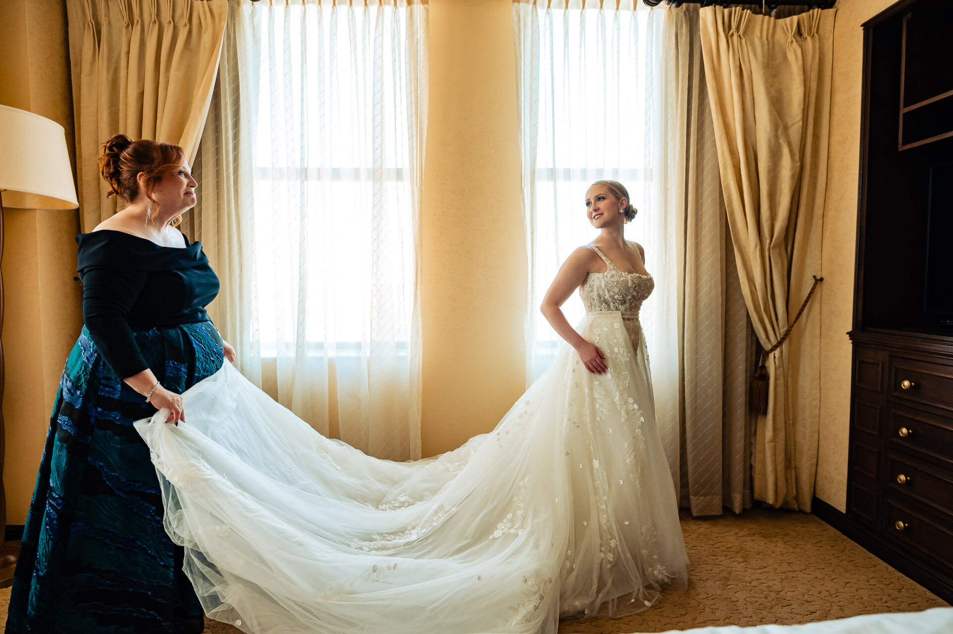 Bride smiling as she adjusts her wedding dress with the help of a bridesmaid in a softly lit room