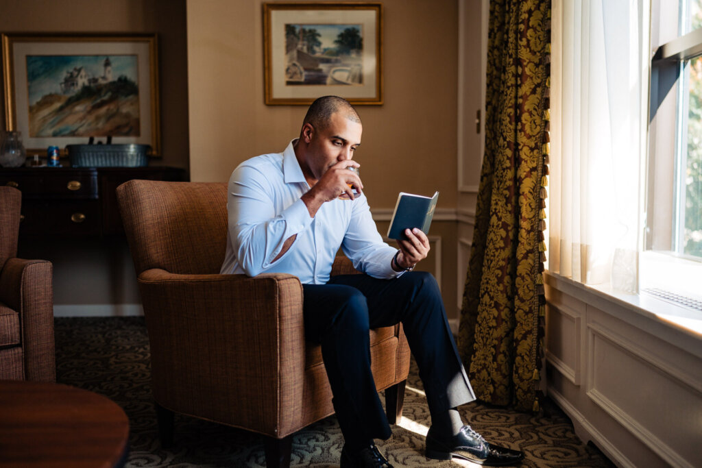 Groom sitting by a window, holding a wedding vow booklet in his hands, gazing out thoughtfully, with sunlight streaming through the curtains