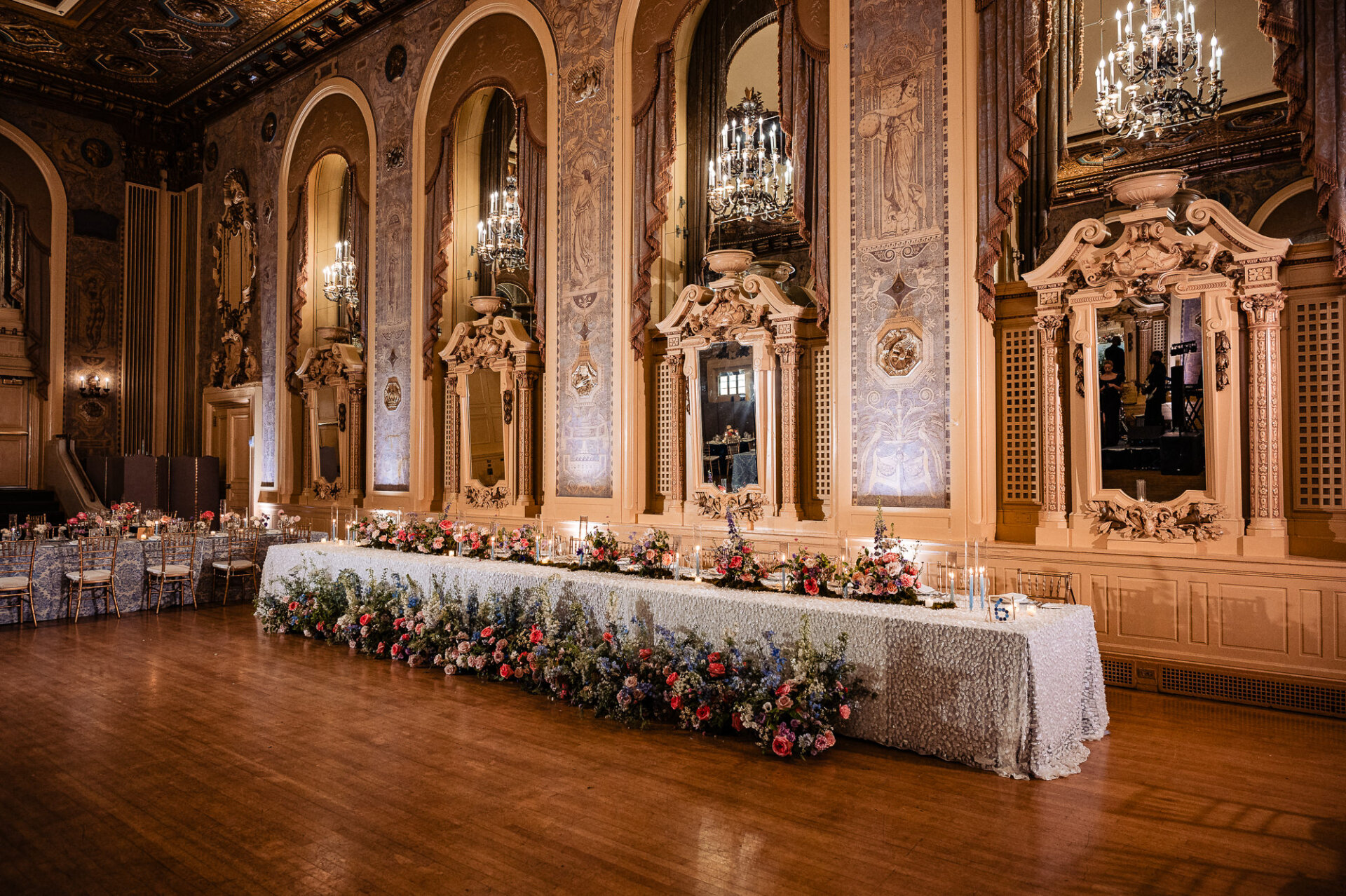 Long wedding reception table set with fine china, crystal glassware, and vibrant floral centerpieces extending the length of the table, with gold chairs and candlelight