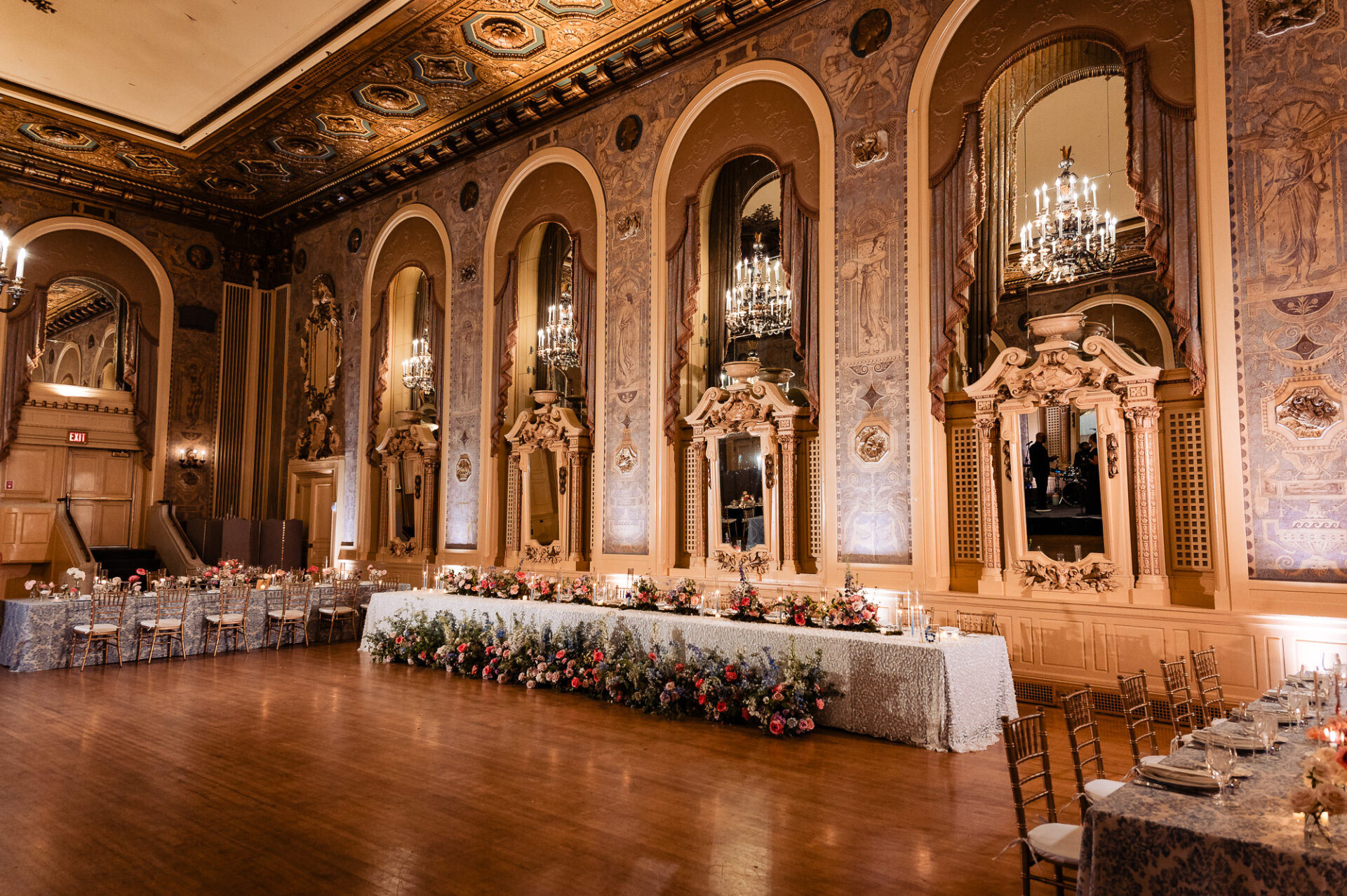 Wide-angle view of the wedding reception hall featuring a grand head table with cascading floral arrangements, chandeliers, and intricate architectural details
