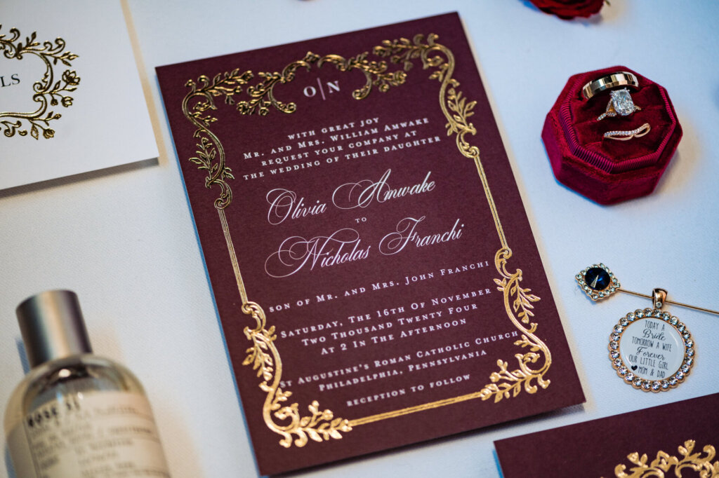Detailed flat lay of a wedding invitation suite with gold floral borders and burgundy background. The scene includes a silver pocket watch, a perfume bottle, and a red rose, all arranged on white fabric with soft lighting emphasizing the textures and colors.