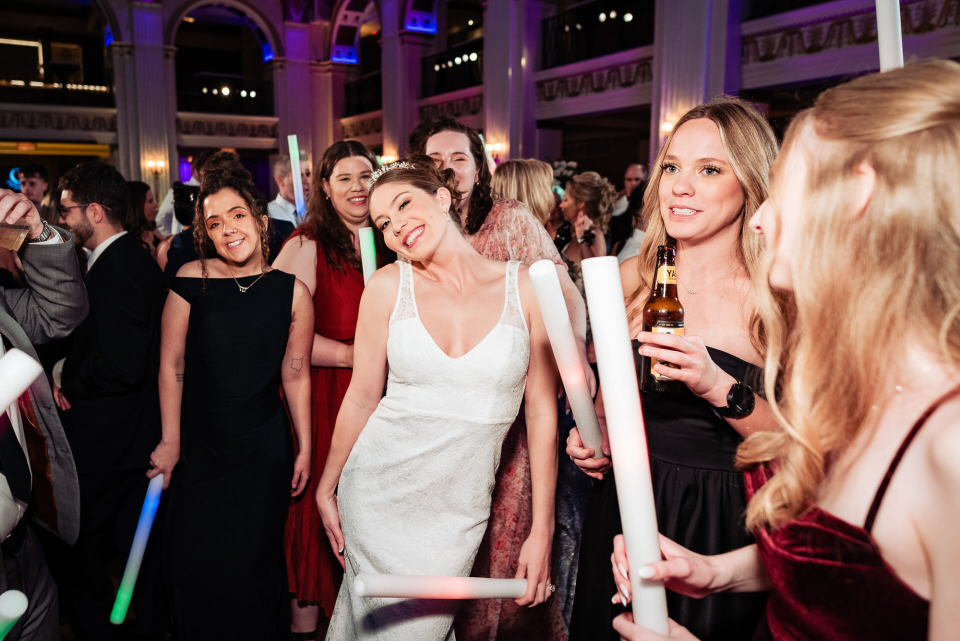 Bride dancing with guests on the dance floor, holding a light-up foam stick and laughing. Guests around her, dressed in formal attire, celebrate with drinks and glow sticks, adding to the lively atmosphere.