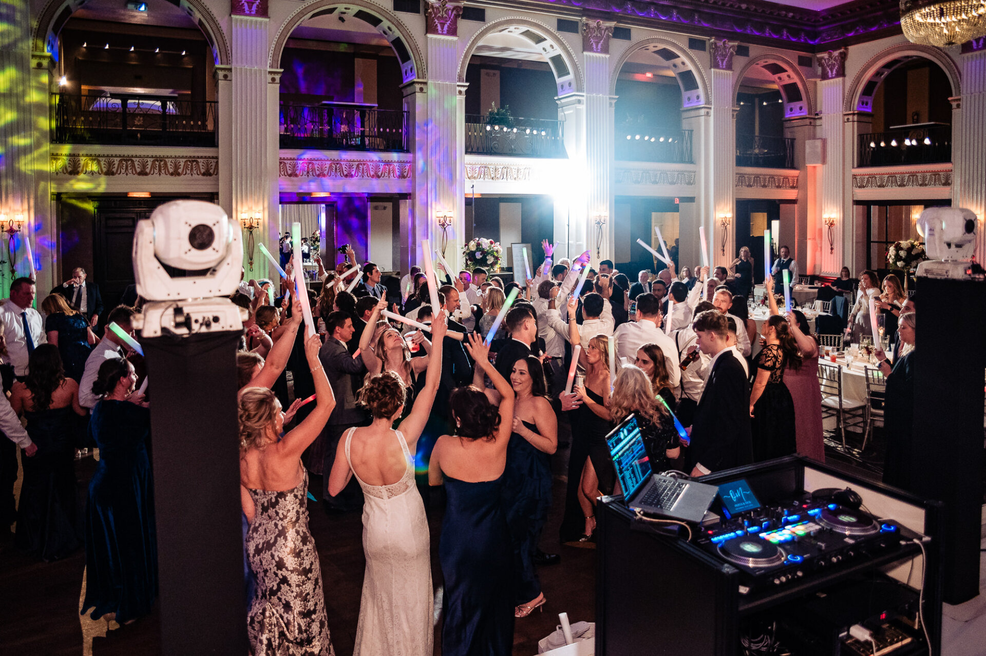 Wedding guests, including the bride, raising glow sticks and dancing together in the reception hall. The DJ booth and colorful uplighting set the scene, while chandeliers and elegant columns frame the background at the Sophisticated Wedding at Ballroom at the Ben reception