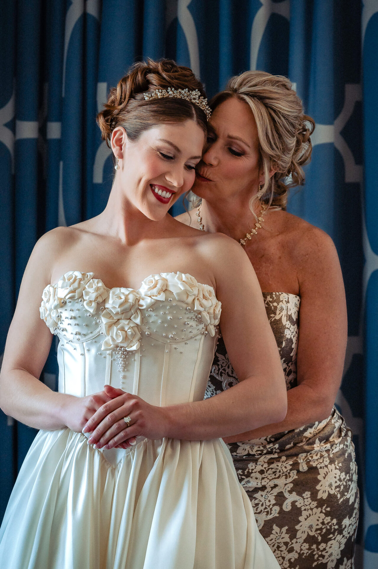 Bride standing with her mother, who embraces her from behind and kisses her on the temple. The bride smiles softly while wearing an ivory gown with floral appliqué details, and the mother wears a lace evening dress.