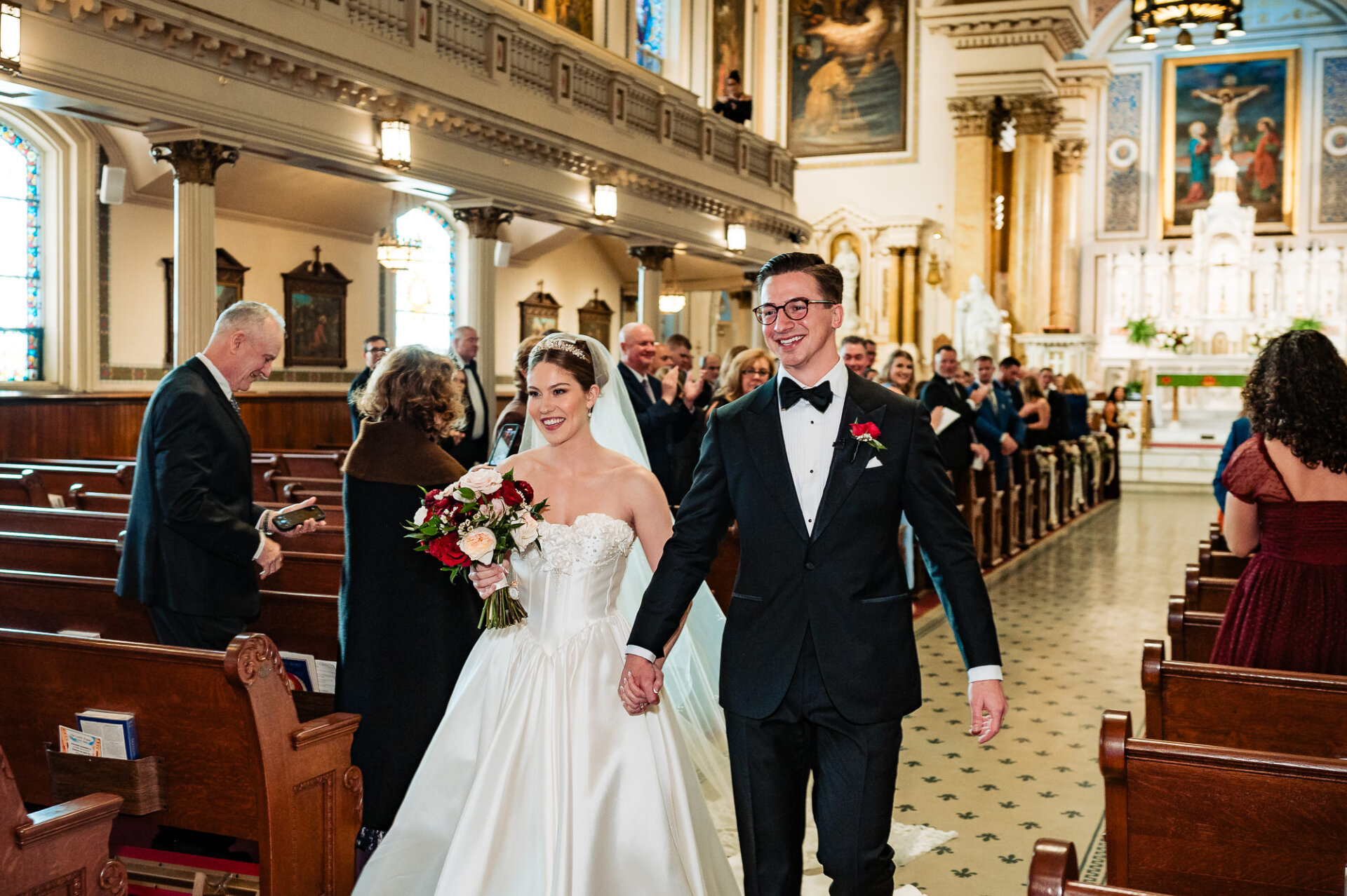 Bride and groom walking down the aisle, hand in hand, after the ceremony. The bride carries a bouquet of red and white flowers, and guests watch as the couple exits the church with smiles.
