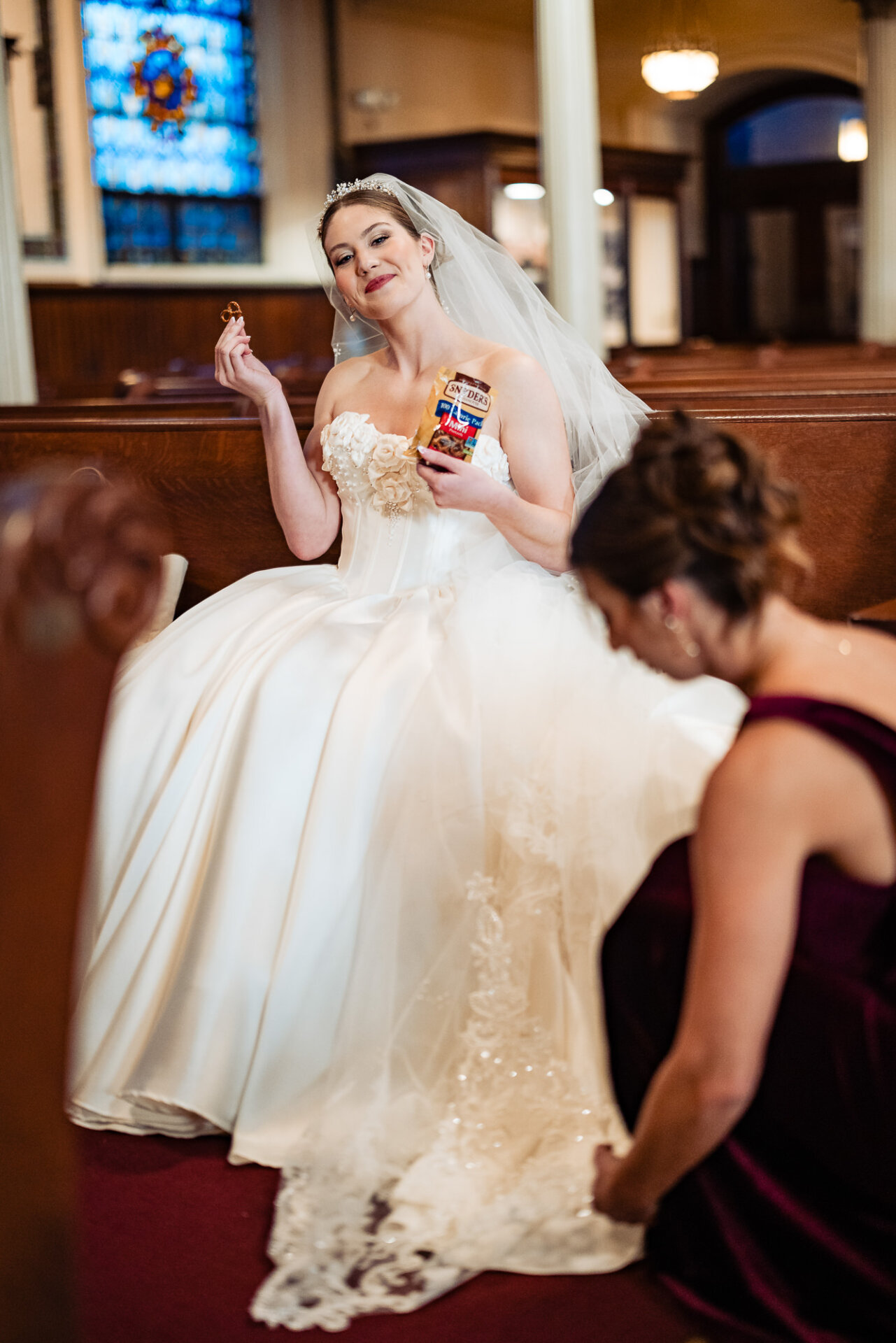 Bridesmaid kneeling to adjust the bride’s gown as the bride sits in the church, lifting her wedding gown slightly to reveal lace details.