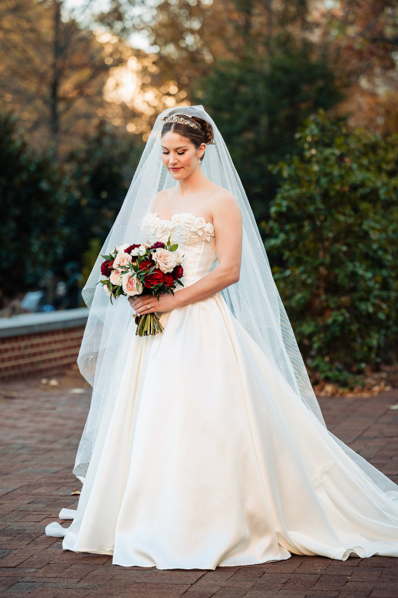 Bride standing on a brick pathway, holding a bouquet of red and blush roses. She smiles softly, with her veil draped over her shoulders and sunlight filtering through the trees behind her.