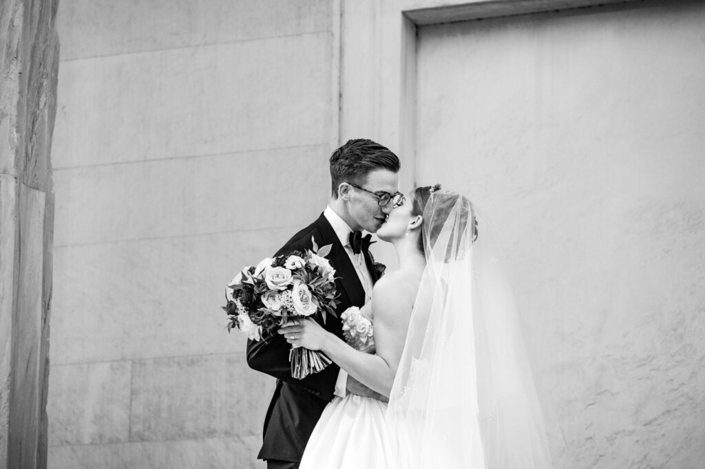 Black and white shot of the groom kissing the bride as she smiles and leans into him. The bride holds a bouquet of roses, and the couple stands against a textured stone wall.