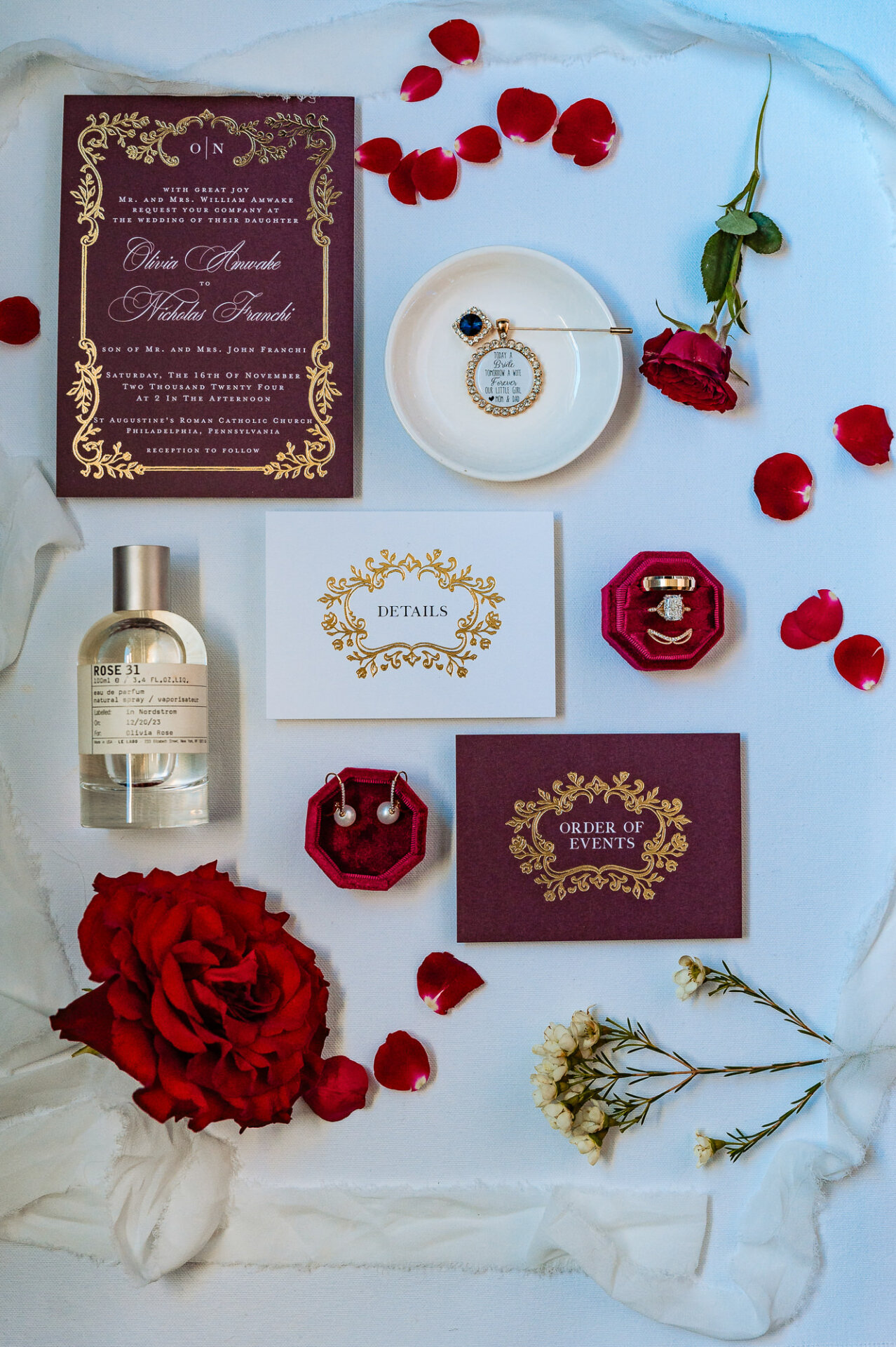 Overhead shot of an elaborate wedding invitation suite featuring a gold-accented burgundy invitation, RSVP card, and wax-sealed envelope. A bottle of perfume, a red velvet ring box with gold trim, a gold pocket watch, and roses are carefully placed around the set on white fabric.